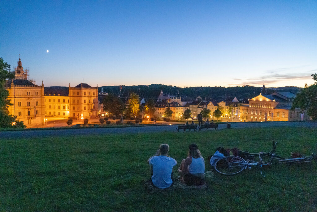 Zwei Personen sitzen in der Abenddämmerung auf einem grasbewachsenen Hügel, in der Nähe liegen Fahrräder, und blicken auf eine von Straßenlaternen beleuchtete Stadtlandschaft, an deren Himmel eine Mondsichel zu sehen ist. at Hochschule Coburg