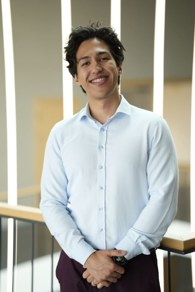 A young man with wavy dark hair, wearing a light blue button-up shirt and a watch, stands indoors smiling, with soft vertical lights and a railing in the background, reflecting his passion for technology and computer science. at Hochschule Coburg