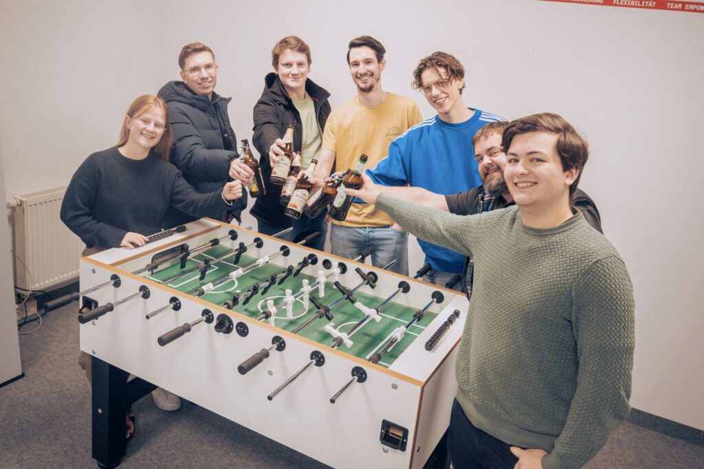 Six young adults stand around a foosball table, smiling and raising bottles in a celebratory toast. The cheerful group enjoys downtime together after an engaging Information Systems 2.0 workshop in a casual indoor setting. at Hochschule Coburg