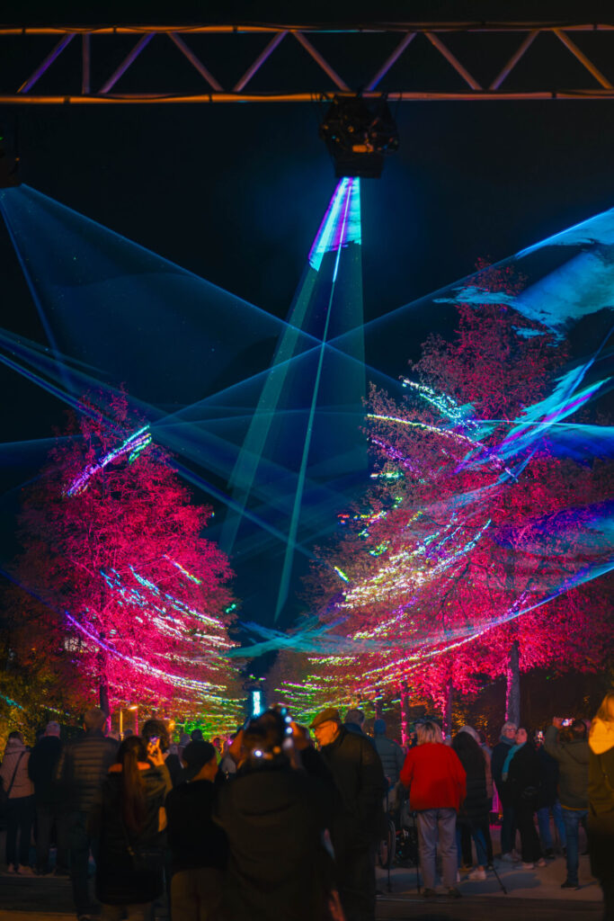 Nachts versammelt sich draußen eine Menschenmenge und beobachtet, wie bunte Lichter und Laserstrahlen die Bäume in leuchtenden Rosa-, Rot- und Blautönen erstrahlen lassen – und so eine festliche, faszinierende Atmosphäre schaffen, die an den Innovationsgeist von „Wirtschaftsinformatik 2.0“ erinnert. at Hochschule Coburg