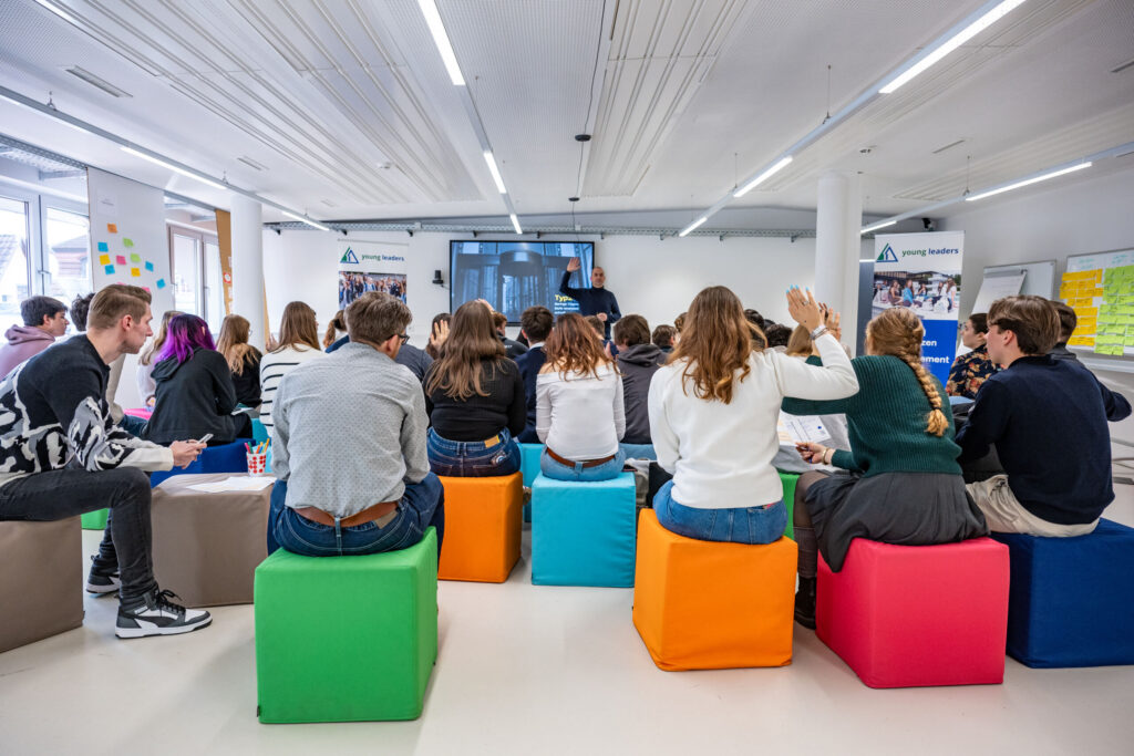A group of young adults sits on colorful cube seats in a bright, modern room, engaging in an Information Systems 2.0 session. Some participants have their hands raised as presentation slides are displayed on the screen behind the speaker. at Hochschule Coburg
