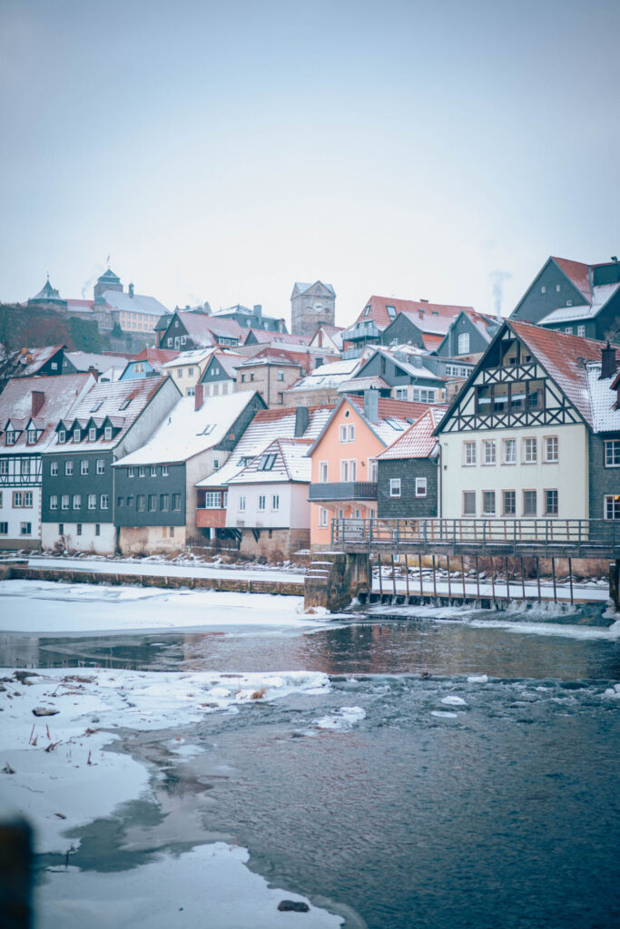 Bunte Häuser mit schneebedeckten Dächern säumen einen Fluss in einer charmanten europäischen Stadt, in der Innovation auf Tradition trifft – so wie die Wirtschaftsinformatik 2.0 unter einer sanften Winterdecke eine Brücke zwischen Vergangenheit und Zukunft schlägt. at Hochschule Coburg