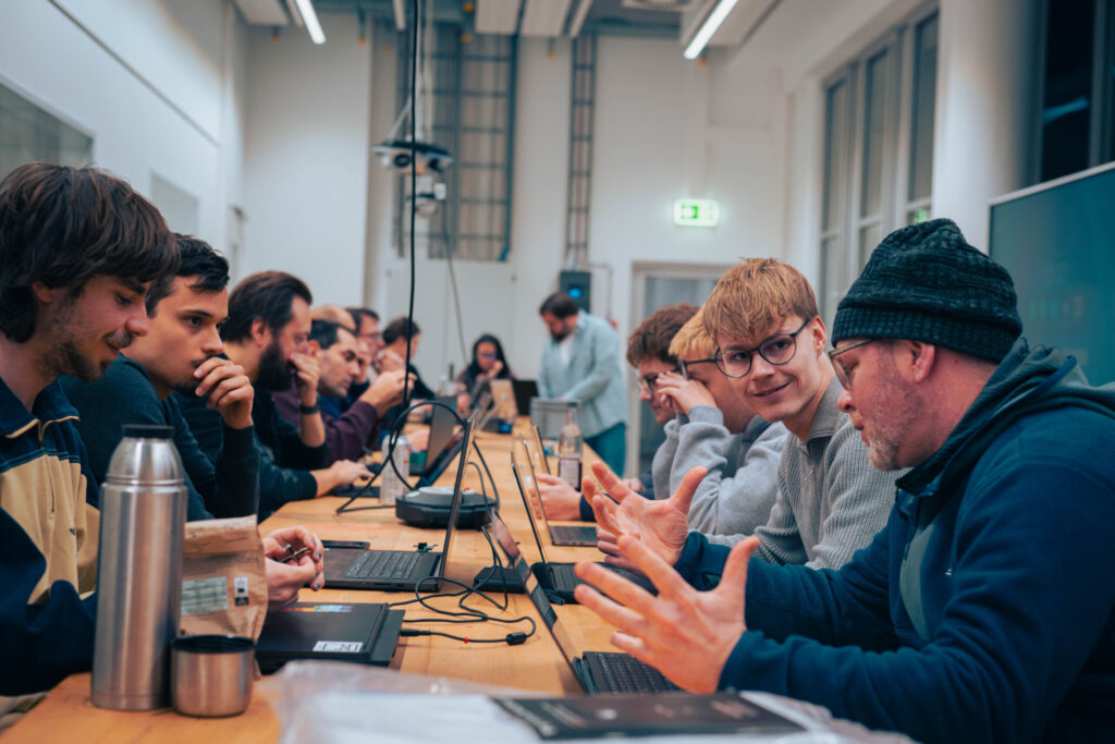 Eine Gruppe von Menschen sitzt mit ihren Laptops an einem langen Tisch und ist in einem hellen, modernen Raum intensiv in Diskussionen und die Zusammenarbeit im Rahmen von „Wirtschaftsinformatik 2.0“ vertieft. Ein Mann gestikuliert lebhaft, während die anderen zuhören und sich an der Diskussion beteiligen. at Hochschule Coburg