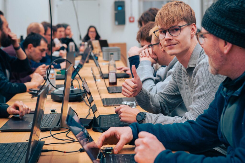 A group of people sit at a long table with laptops, immersed in Information Systems 2.0. One young man in glasses and a gray sweater gives a thumbs up and smiles at the camera, while others focus on their screens in a collaborative atmosphere. at Hochschule Coburg