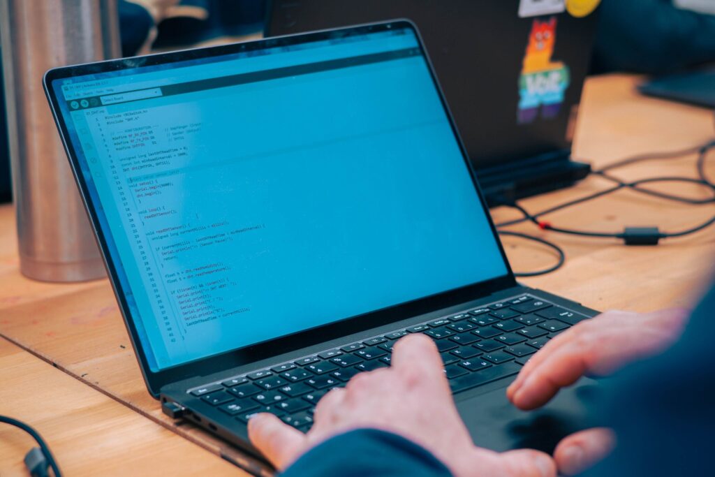 A person is typing on a laptop displaying Information Systems 2.0 code on the screen. Another laptop with colorful stickers and a metal tumbler are visible in the background on a wooden desk. at Hochschule Coburg