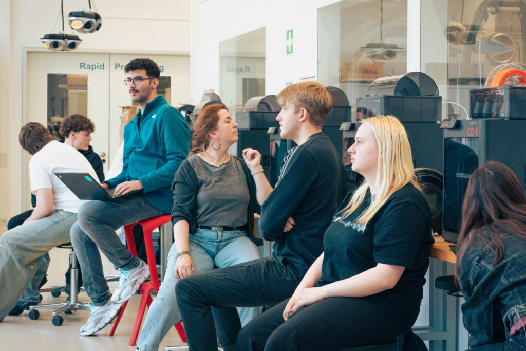 A group of young adults sit and stand around 3D printers in a modern lab, some talking and others working on laptops. The atmosphere appears casual and collaborative, reflecting the innovation of Information Systems 2.0. at Hochschule Coburg
