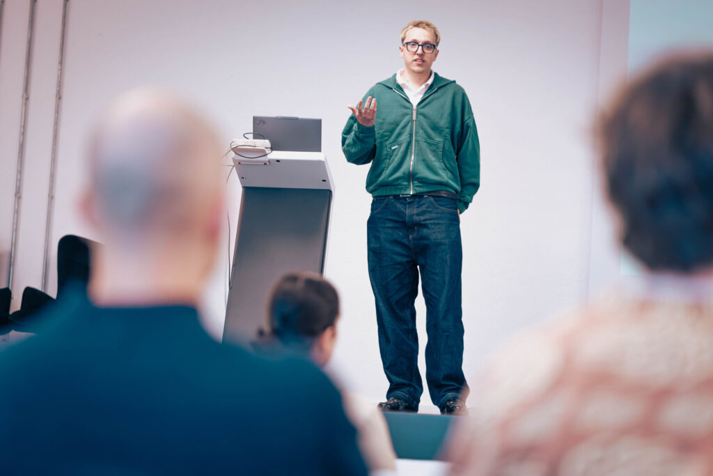 Eine Person in einem grünen Kapuzenpulli und mit Brille steht vor einem Rednerpult und spricht zu den Zuhörern; sie leitet ein Seminar zum Thema „Wirtschaftsinformatik 2.0“. Im Vordergrund des Unterrichtsraums sind mehrere sitzende Teilnehmer zu sehen. at Hochschule Coburg