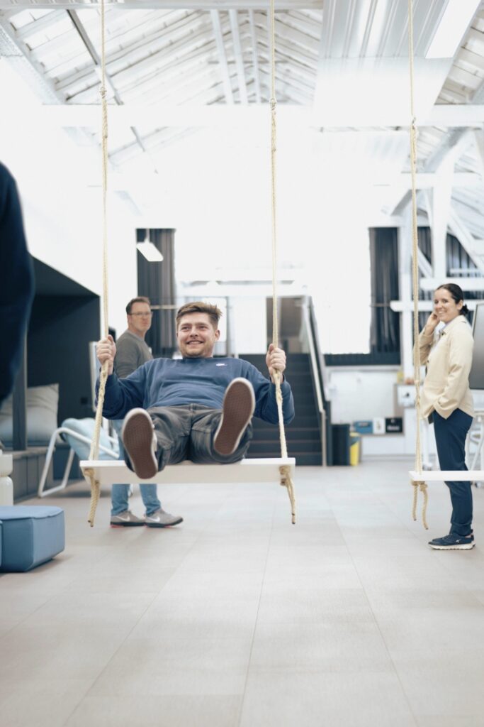 Ein Mann in einem blauen Pullover lächelt, während er auf einer Innenschaukel schaukelt; im Hintergrund eines modernen, hellen Raums, der vom Konzept der Wirtschaftsinformatik 2.0 inspiriert ist, stehen zwei Personen. at Hochschule Coburg