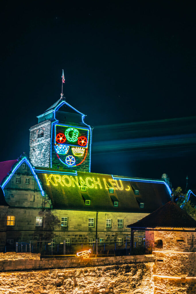 Ein historisches Steingebäude, das nachts von bunten Neonlichtern beleuchtet wird, die sein Dach und seine Fenster umrahmen; ein leuchtendes Wappen und die Aufschrift „KRONACH LEUCHTET“ sind an der Fassade gut sichtbar angebracht und verkörpern den Innovationsgeist der Wirtschaftsinformatik 2.0. at Hochschule Coburg