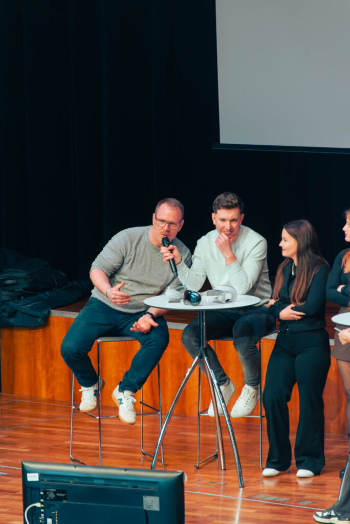 Two men sit on high stools at a round table on stage, one speaking into a microphone. Two women stand nearby, one facing the audience. A large monitor and projector display Information Systems 2.0 in the foreground. at Hochschule Coburg
