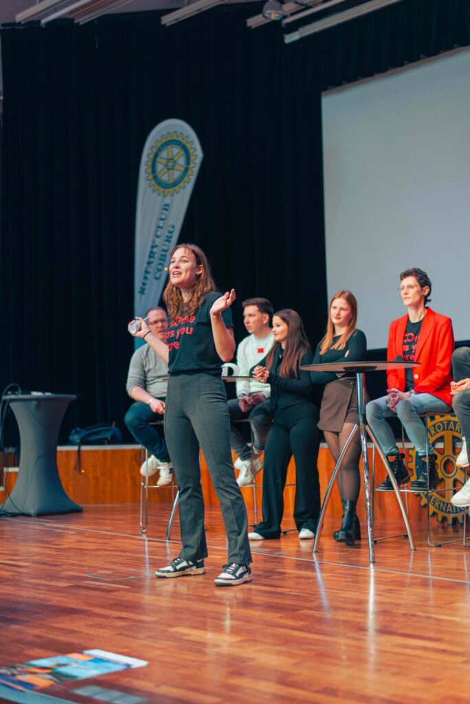 A woman speaks on stage with expressive gestures about Information Systems 2.0, while four people sit behind her at a panel. A Rotary Club banner and a large screen are visible in the background, suggesting a conference or public event. at Hochschule Coburg