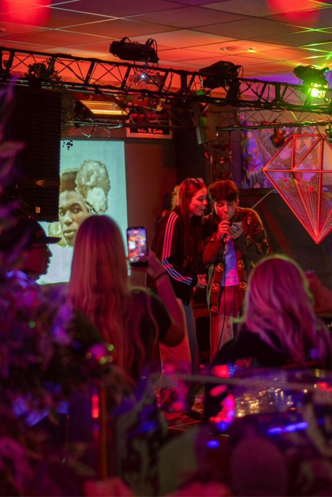 Zwei Personen singen auf einer farbenfrohen, neonbeleuchteten Bühne in ein Mikrofon, während jemand sie mit einem Handy filmt. Hinter ihnen werden ein Schwarz-Weiß-Bild und die Worte „Wirtschaftsinformatik 2.0“ projiziert, während andere aus dem Vordergrund zuschauen. at Hochschule Coburg