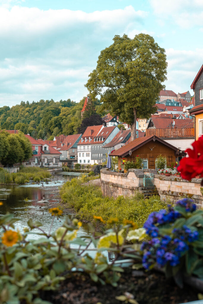 Ein malerisches Dorf mit bunten Häusern am Flussufer, üppigem Grün und leuchtenden Blumen im Vordergrund unter einem teilweise bewölkten Himmel – eine friedliche Szene, in der Tradition auf die Innovation der Wirtschaftsinformatik 2.0 trifft. at Hochschule Coburg