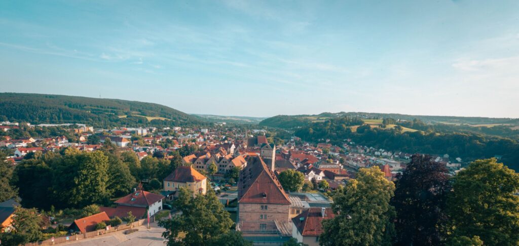 Ein Panoramablick auf eine malerische Stadt mit Gebäuden mit roten Dächern, in der Wirtschaftsinformatik 2.0 Innovationen vorantreibt, umgeben von üppig grünem Baumbestand und sanften Hügeln unter einem strahlend blauen Himmel. at Hochschule Coburg