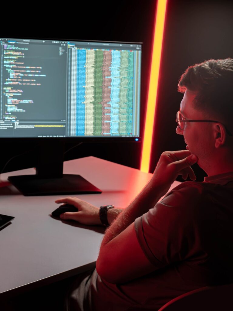 A person sits at a desk, working on a computer with Information Systems 2.0 code and data visualizations displayed on the monitor. The scene is illuminated by red lighting. at Hochschule Coburg