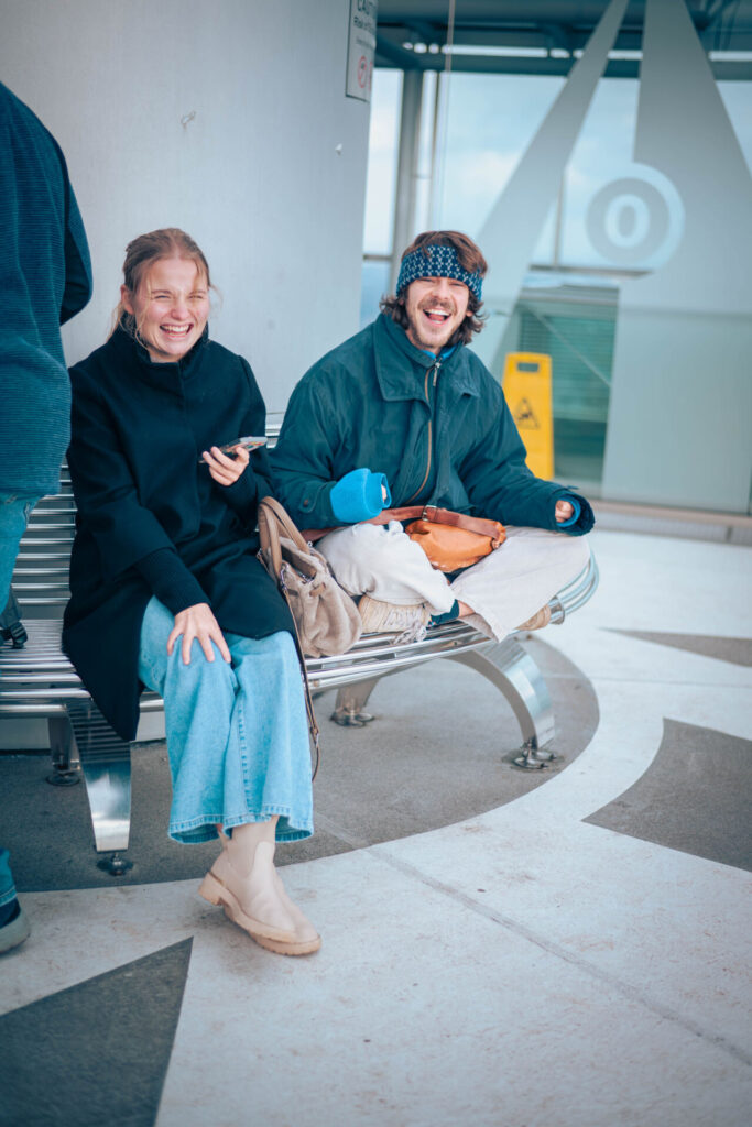 Zwei Personen sitzen drinnen auf einer Metallbank, lächeln und lachen. Die eine trägt einen dunklen Mantel und Jeans, die andere eine blaue Jacke und ein Stirnband und hält einen blauen Fäustling in der Hand – vielleicht unterhalten sie sich gerade über Wirtschaftsinformatik 2.0. Neben ihnen liegen Taschen auf der Bank. at Hochschule Coburg