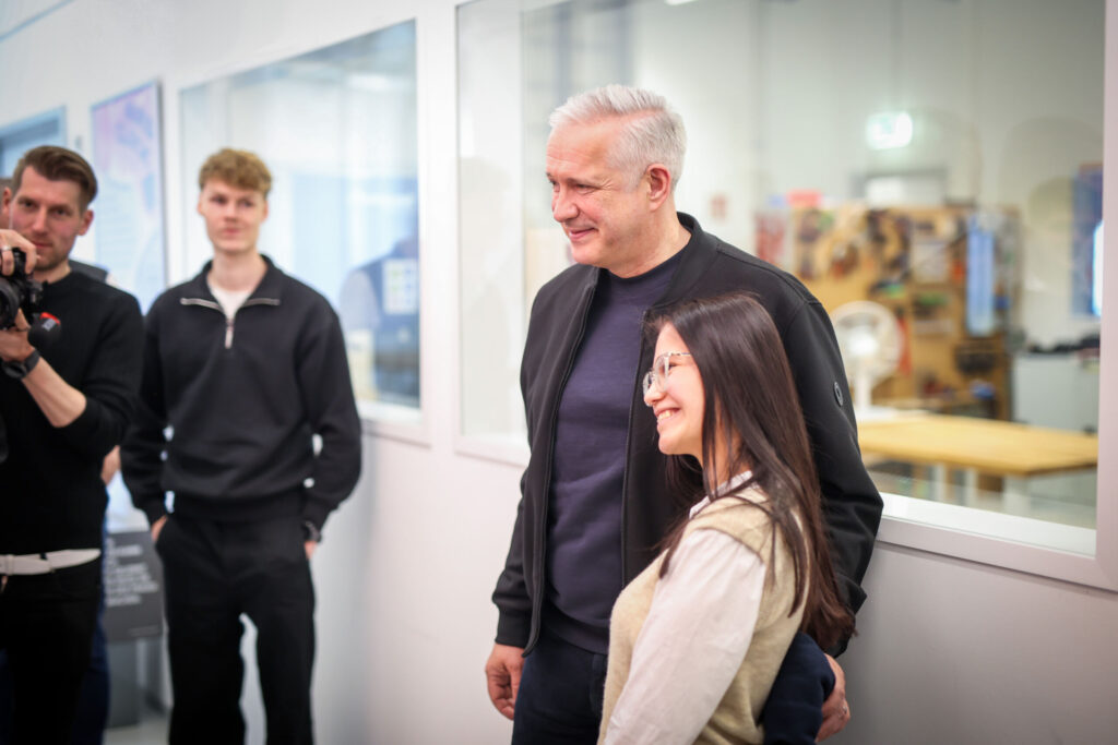 A man and a woman stand together smiling for a photo indoors at an Information Systems 2.0 event, while others watch in the background. The modern setting features large windows and bright lighting. at Hochschule Coburg