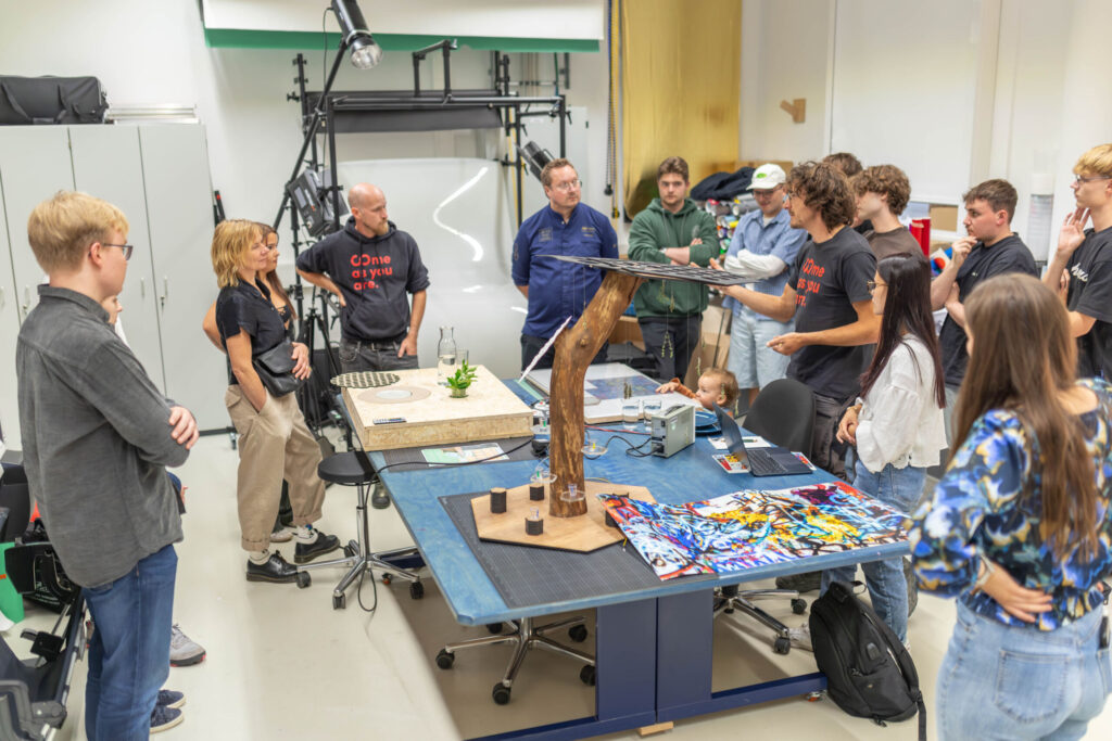 A group of people stand around a table covered with artwork and models in a bright studio, engaged in discussion. One person appears to be presenting or explaining an object, highlighting concepts inspired by Information Systems 2.0. at Hochschule Coburg