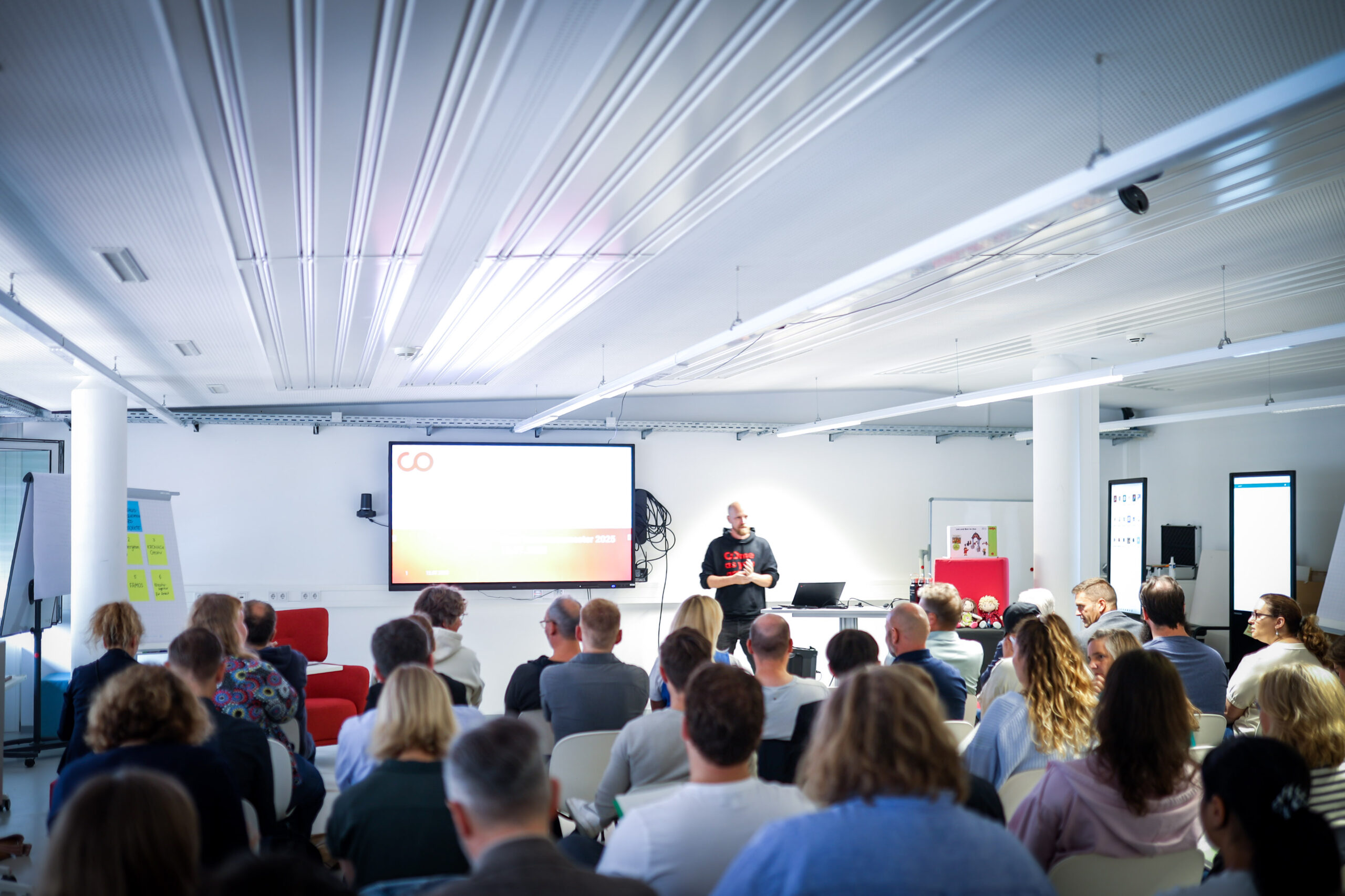In einem modernen, hell erleuchteten Raum in Kronach sitzt eine Gruppe von Menschen einem Referenten gegenüber. Der Referent steht neben einer großen Leinwand, auf der eine ZukunftsDesign-Präsentation zu sehen ist, während das Publikum diesem spannenden Experiment aufmerksam lauscht. at Hochschule Coburg