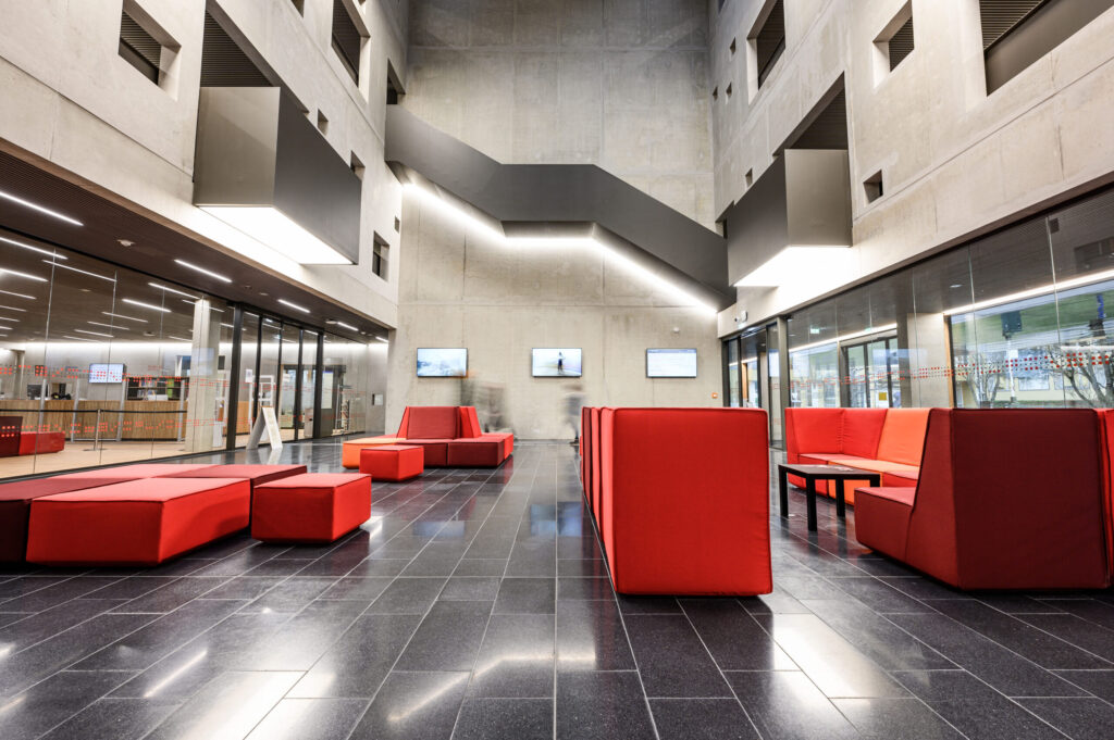 Moderne, geräumige Lobby mit grauen Böden, roten Sitzpolstern, hohen Betonwänden und großen Fenstern. Drei an der Wand befestigte Bildschirme und eine Treppe sind sichtbar und schaffen eine klare, moderne Atmosphäre. at Hochschule Coburg
