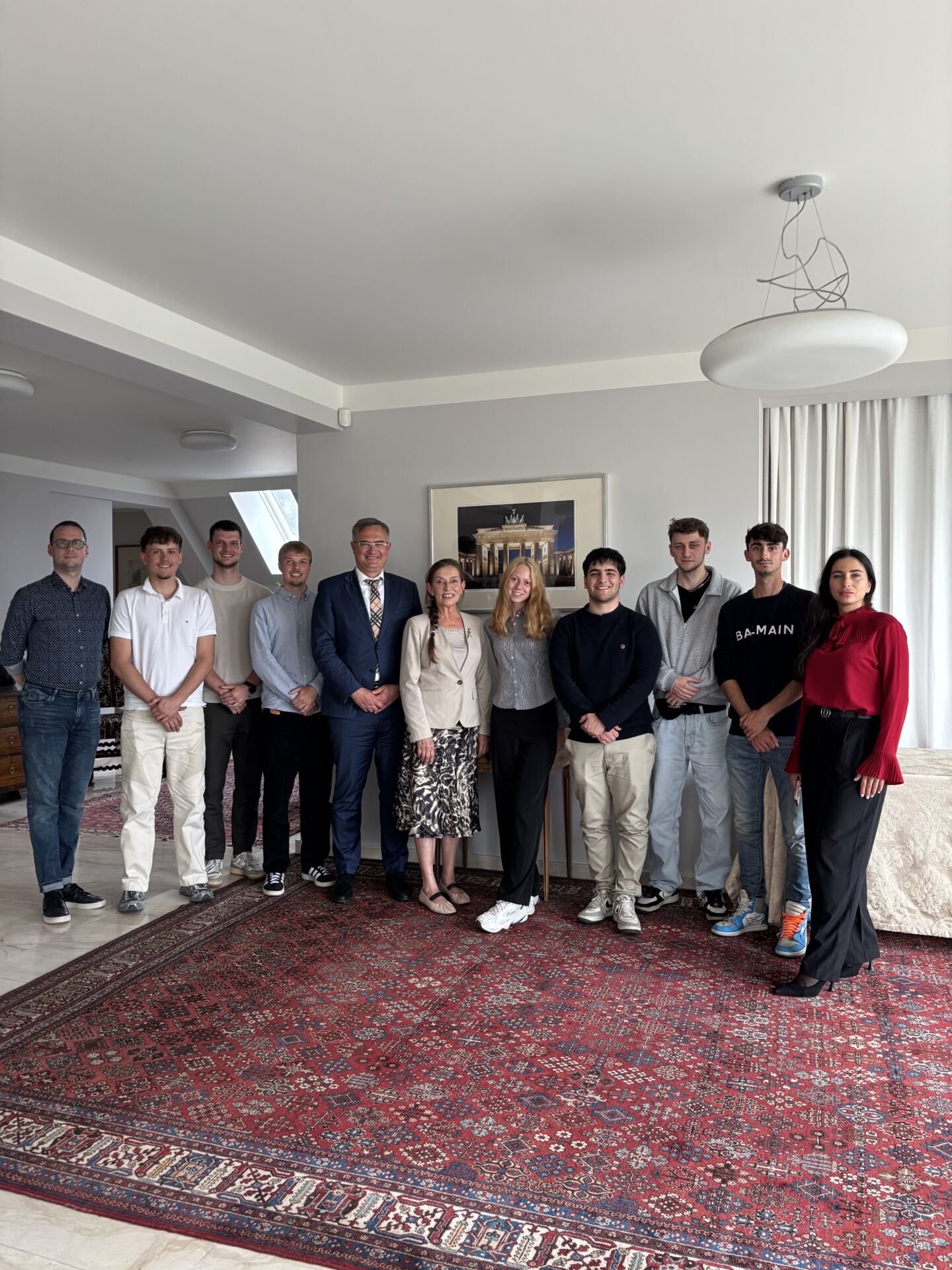 Eine Gruppe von zehn Personen, gekleidet in Business-Casual- und Freizeitkleidung, steht in einem gut beleuchteten Raum auf einem gemusterten roten Teppich. Sie posieren und lächeln für die Kamera vor einem gerahmten Bild, neben dem ein automatisch gespeicherter Entwurf liegt. at Hochschule Coburg