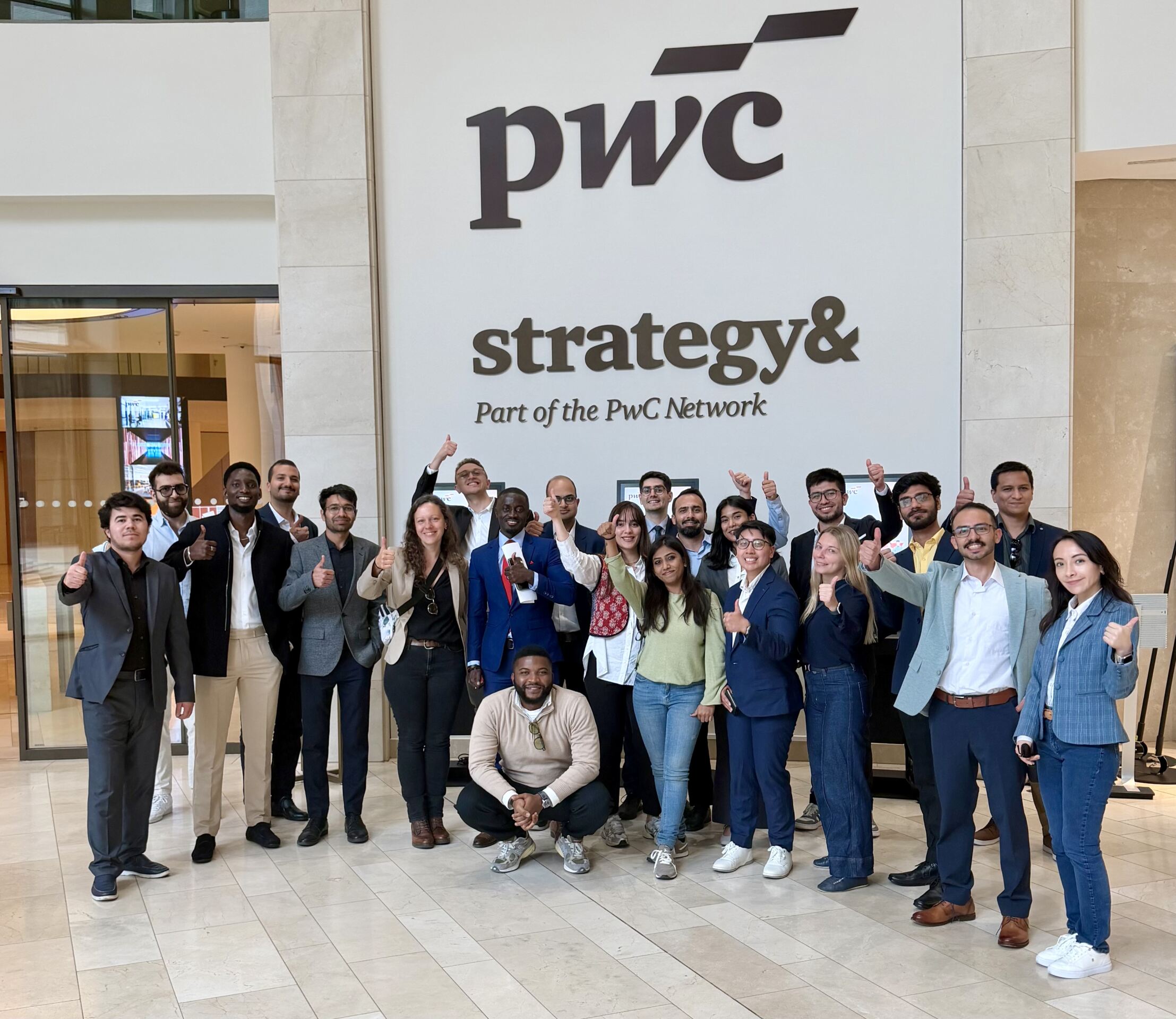 Eine bunt gemischte Gruppe von Menschen posiert lächelnd und mit erhobenem Daumen vor einem großen „PwC strategy&“-Schild in einem modernen Bürogebäude. at Hochschule Coburg