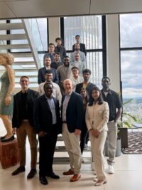 A group of sixteen professionally dressed people posing and smiling on a modern indoor staircase, with large windows and a cityscape visible in the background. A sculpture stands on the left side of the image. at Hochschule Coburg