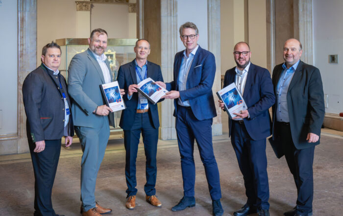Sechs Männer in Geschäftskleidung stehen in einem Haus und posieren für ein Foto. Drei von ihnen zeigen einen Automatisch gespeicherten Entwurf, während die anderen lächelnd daneben stehen. Der formelle Rahmen besteht aus Marmorsäulen und gewölbten Türöffnungen. at Hochschule Coburg