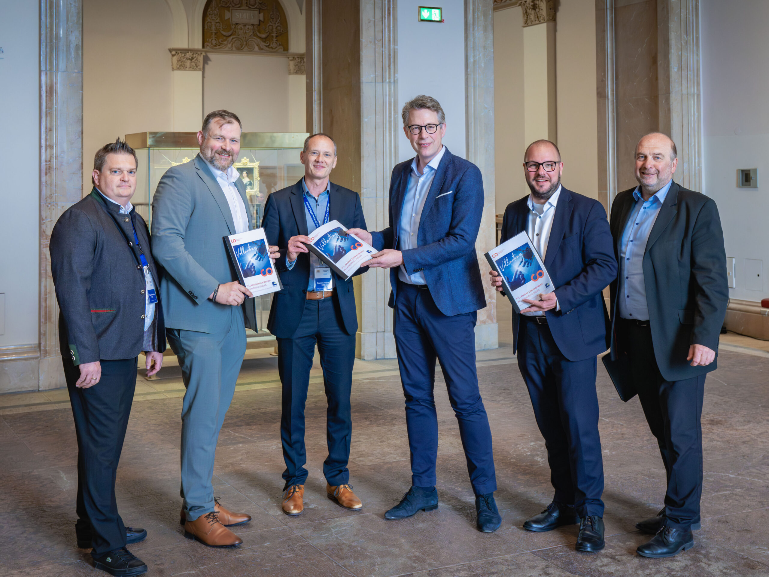 Sechs Männer in Geschäftskleidung stehen in einem Haus und posieren für ein Foto. Drei von ihnen zeigen einen Automatisch gespeicherten Entwurf, während die anderen lächelnd daneben stehen. Der formelle Rahmen besteht aus Marmorsäulen und gewölbten Türöffnungen. at Hochschule Coburg