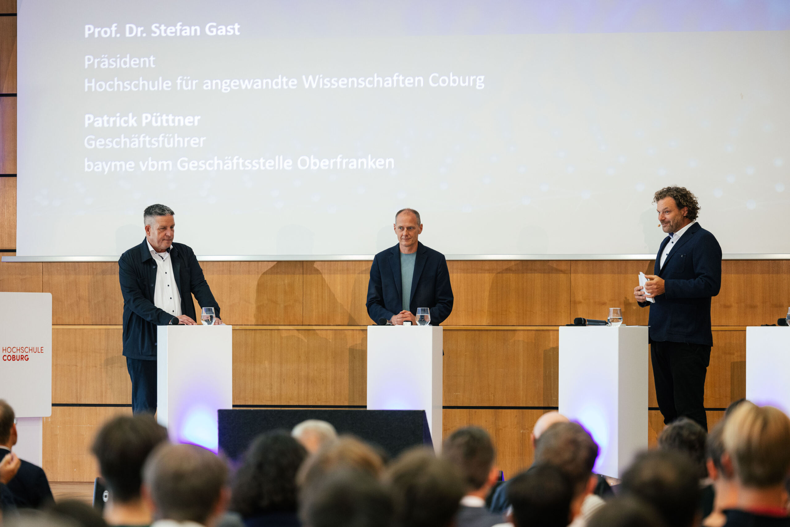 Drei Männer in Geschäftskleidung stehen hinter separaten Rednerpult auf der Bühne und beteiligen sich an der Initiative „Chancen durch Dialog“, um Wissenschaft und Industrie miteinander zu verbinden. Auf der großen Leinwand hinter ihnen werden bei dieser formellen Konferenz ihre Namen und Titel auf Deutsch angezeigt. at Hochschule Coburg