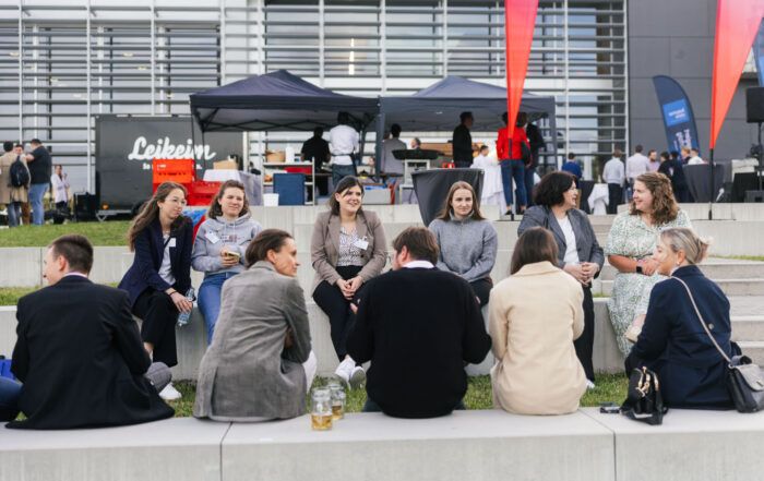 Eine Gruppe von Menschen sitzt im Freien auf Steinbänken im Kreis, unterhält sich und lächelt bei der Veranstaltung „Dialog Wissenschaft und Wirtschaft“, vor dem Hintergrund von Imbissständen, den „3x3 Brücken“-Installationen und einem modernen Gebäude. at Hochschule Coburg