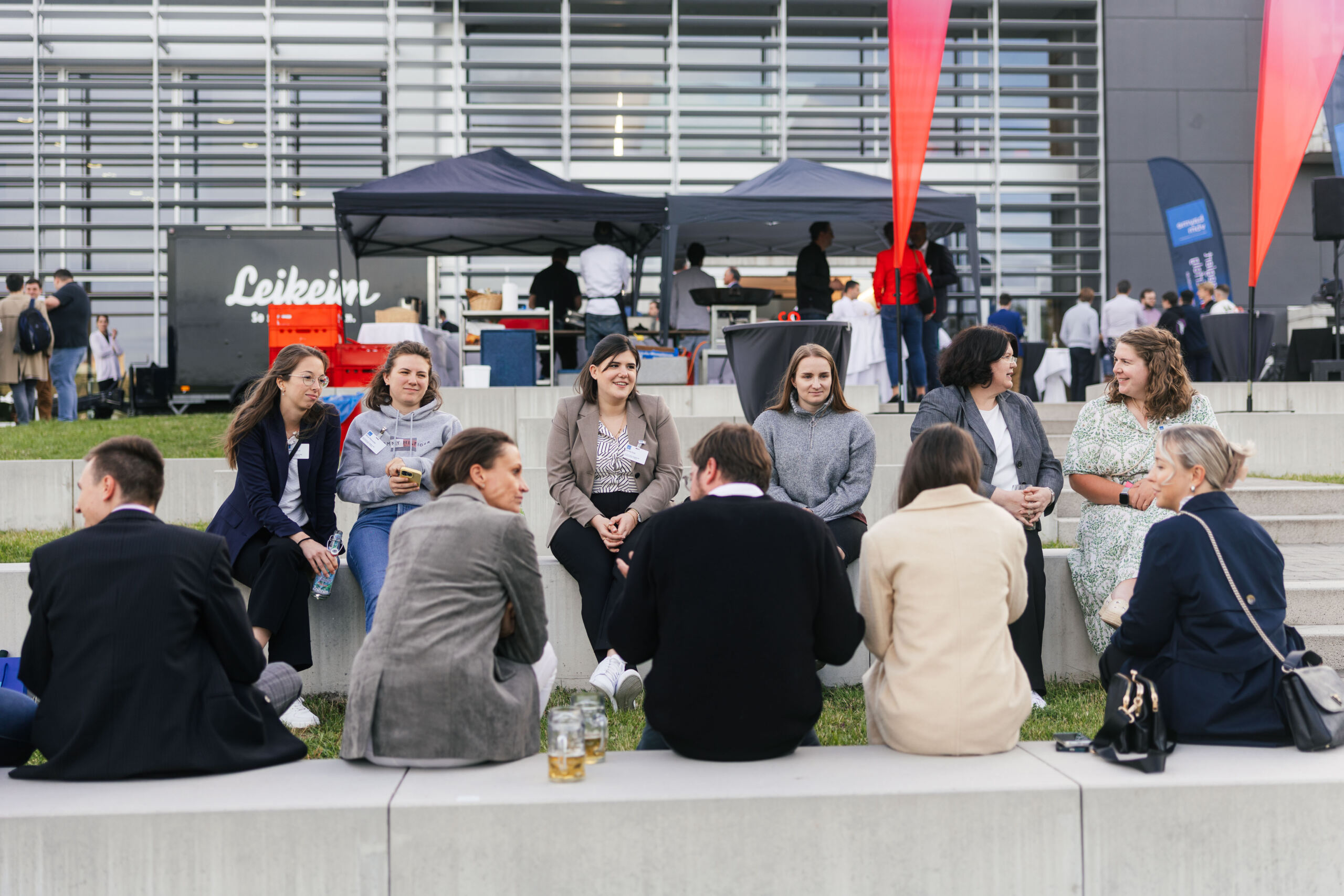 Eine Gruppe von Menschen sitzt im Freien auf Steinbänken im Kreis, unterhält sich und lächelt bei der Veranstaltung „Dialog Wissenschaft und Wirtschaft“, vor dem Hintergrund von Imbissständen, den „3x3 Brücken“-Installationen und einem modernen Gebäude. at Hochschule Coburg