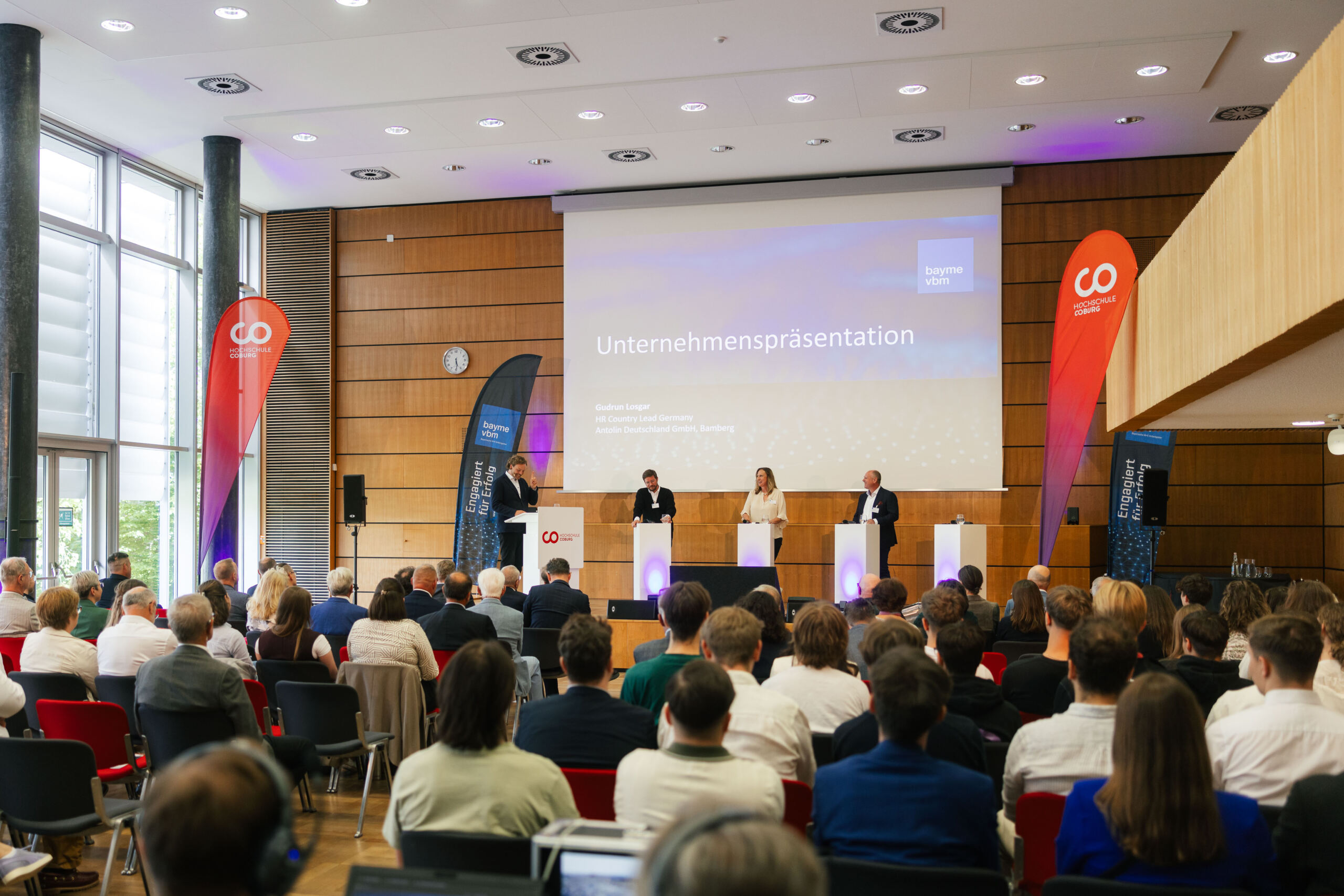 Eine Unternehmenspräsentation in einem modernen Auditorium, bei der vier Redner auf der Bühne vor einem aufmerksamen Publikum einen Dialog führen. Große Banner und eine Leinwand mit der Aufschrift „Unternehmenspräsentation“ unterstreichen den Schwerpunkt auf Wirtschaft und Innovation. at Hochschule Coburg