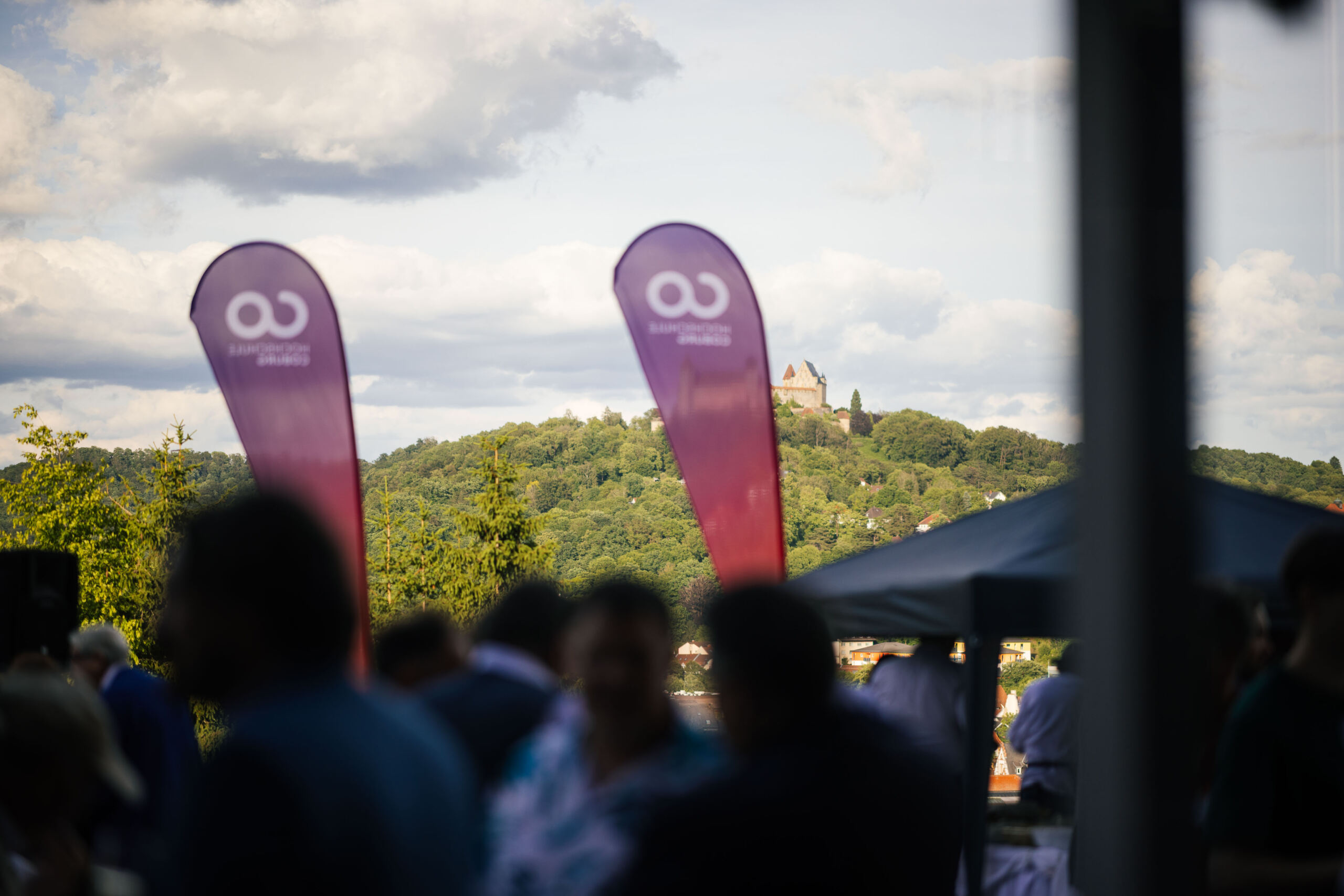 Im Vordergrund sind Menschen im Freien zu sehen, die zwei violette Fahnen in der Hand halten und sich inmitten einer verschwommenen Menschenmenge unterhalten; im Hintergrund sind unter einem teilweise bewölkten Himmel ein grüner Hügel und eine Burg darauf zu erkennen. at Hochschule Coburg