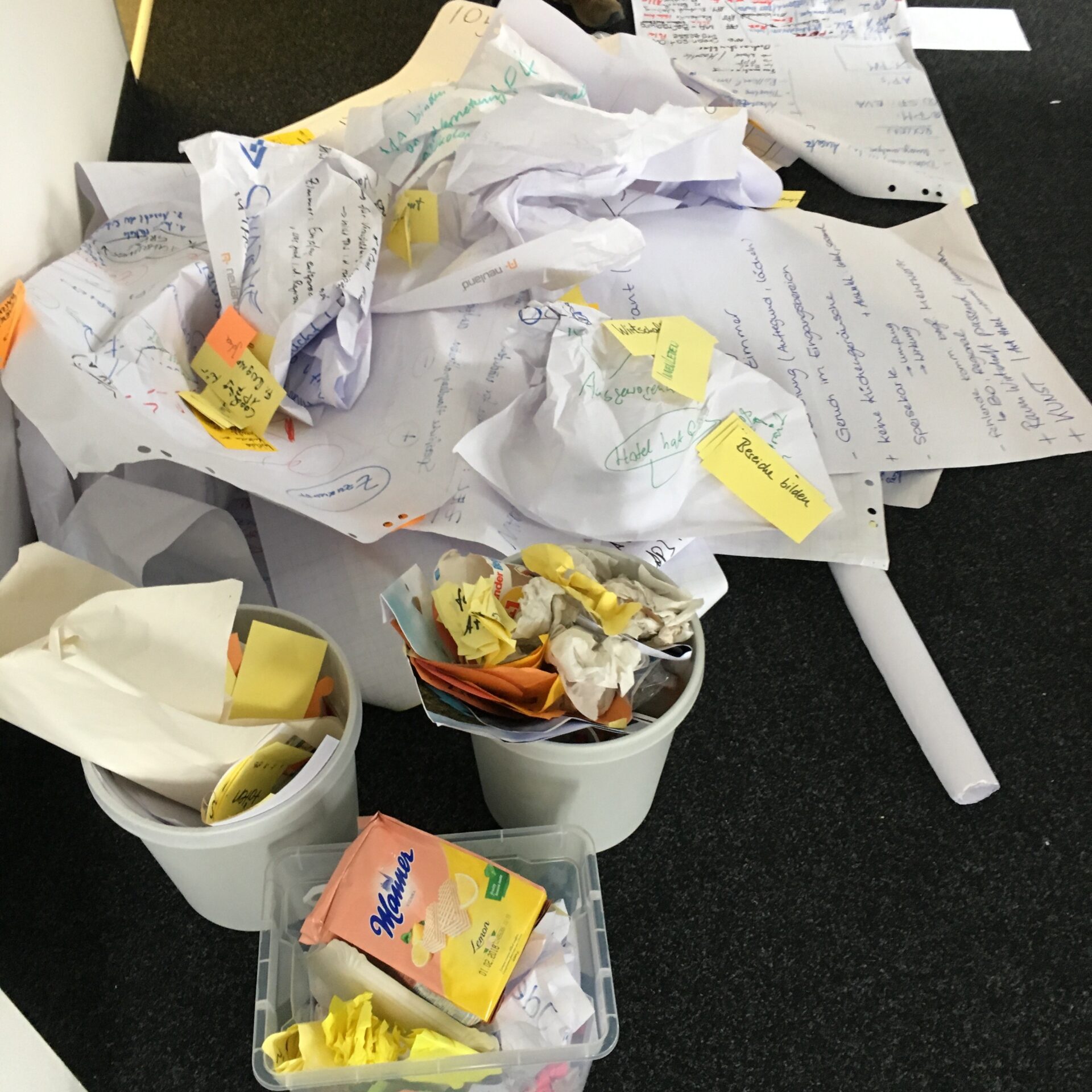 Zerknüllte Zettel, Haftnotizen und weggeworfene Notizen liegen verstreut auf dem Boden und stapeln sich in Behältern – ein Bild, das einen unaufgeräumten Arbeitsplatz widerspiegelt, an dem Experimentierfreude auf kreative Arbeit trifft – vielleicht während einer ZukunftsDesign-Sitzung in Kronach. at Hochschule Coburg