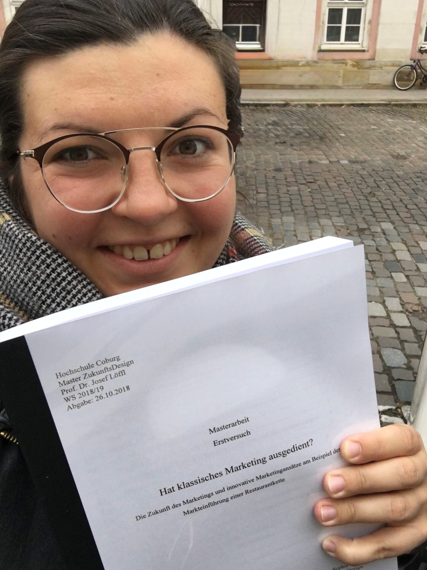 Eine lächelnde Person mit Brille und Schal hält eine gedruckte Abhandlung mit dem Titel „Hat klassisches Marketing ausgedient?“ – ein Experiment in Zukunftsdesign – in die Höhe, während sie draußen auf einer Kopfsteinpflasterstraße in Kronach vor einem Gebäude mit weißen Fenstern steht. at Hochschule Coburg