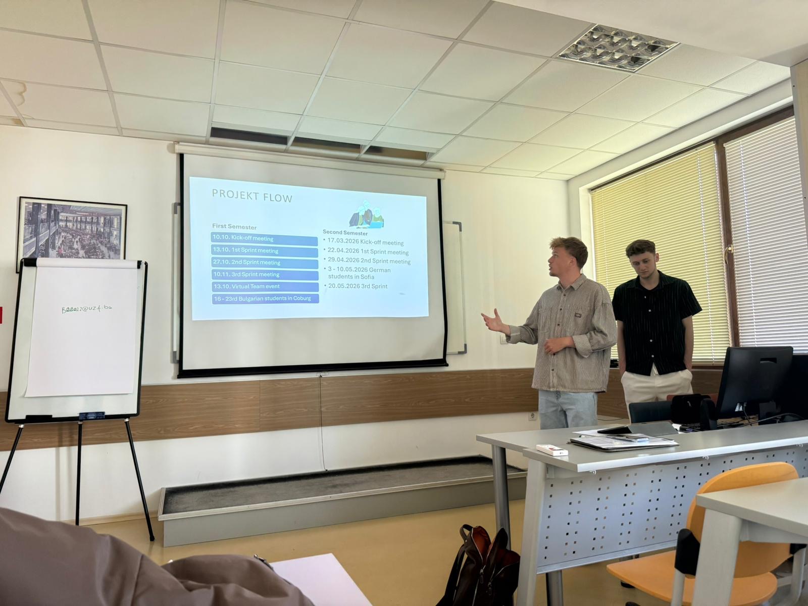 Zwei Personen stehen vorne im Klassenzimmer und halten einen Vortrag. Hinter ihnen wird eine Folie mit dem Titel „Projektablauf“ projiziert, die als „ein automatisch gespeicherter Entwurf“ gekennzeichnet ist, während eine der Personen darauf zeigt. Man sieht Tische, Stühle und eine Tafel. at Hochschule Coburg
