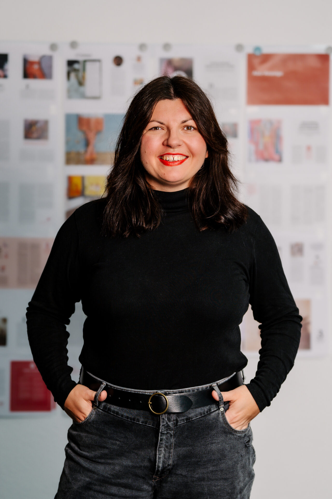 Eine Frau mit dunklem Haar, die einen schwarzen Rollkragenpullover und schwarze Jeans trägt, steht lächelnd mit den Händen in den Taschen vor einer Wand, die mit bunten Papieren und Bildern bedeckt ist, beim ZukunftsDesign Experiment in Kronach. at Hochschule Coburg