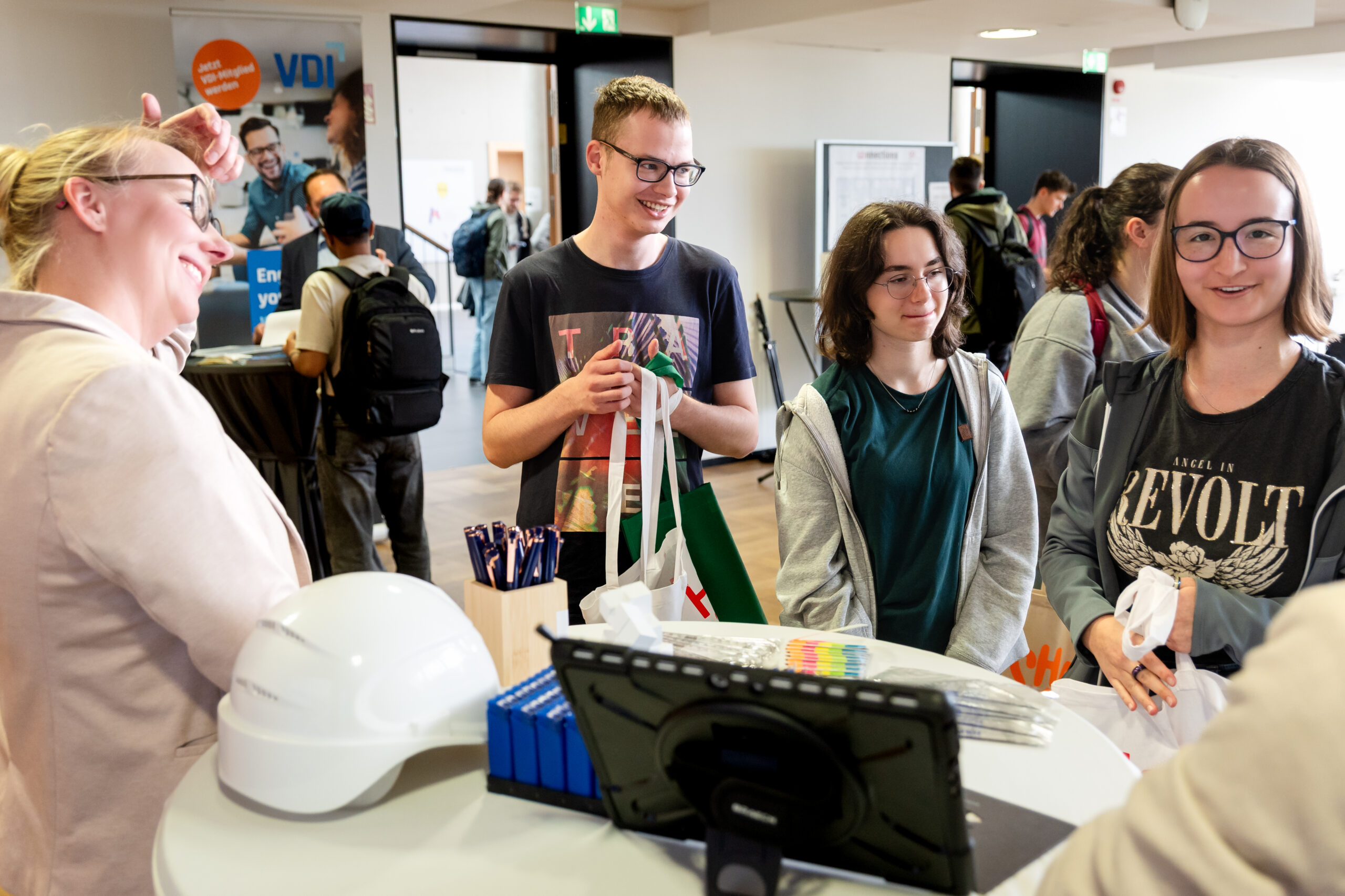 Vier junge Erwachsene stehen an einem Stand und unterhalten sich mit einer Frau, die wertvolle Kontakte knüpft. Auf dem Tisch liegen Werbeartikel, darunter Stifte und ein Schutzhelm. Die lockere Atmosphäre ist perfekt, um unter den sichtbaren Teilnehmern im Hintergrund Talente zu gewinnen. at Hochschule Coburg
