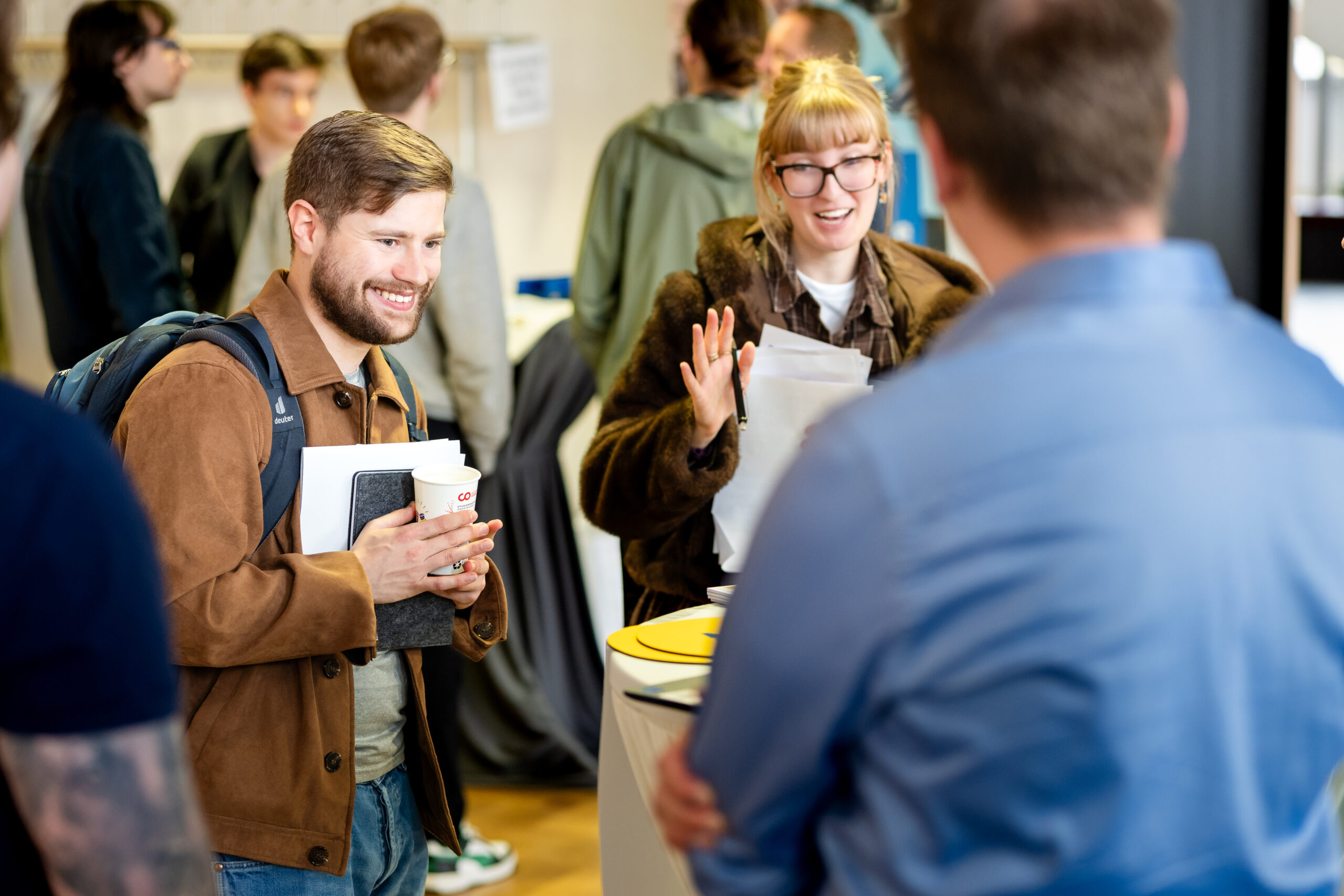 Ein Mann mit einem Kaffee und einem Notizbuch in der Hand lächelt, während er sich mit einer Frau unterhält, die bei einer belebten Indoor-Veranstaltung Papiere in der Hand hält - ein ideales Umfeld, um Talente zu gewinnen und wertvolle Kontakte zu knüpfen, während sich andere im Hintergrund mischen und unterhalten. at Hochschule Coburg