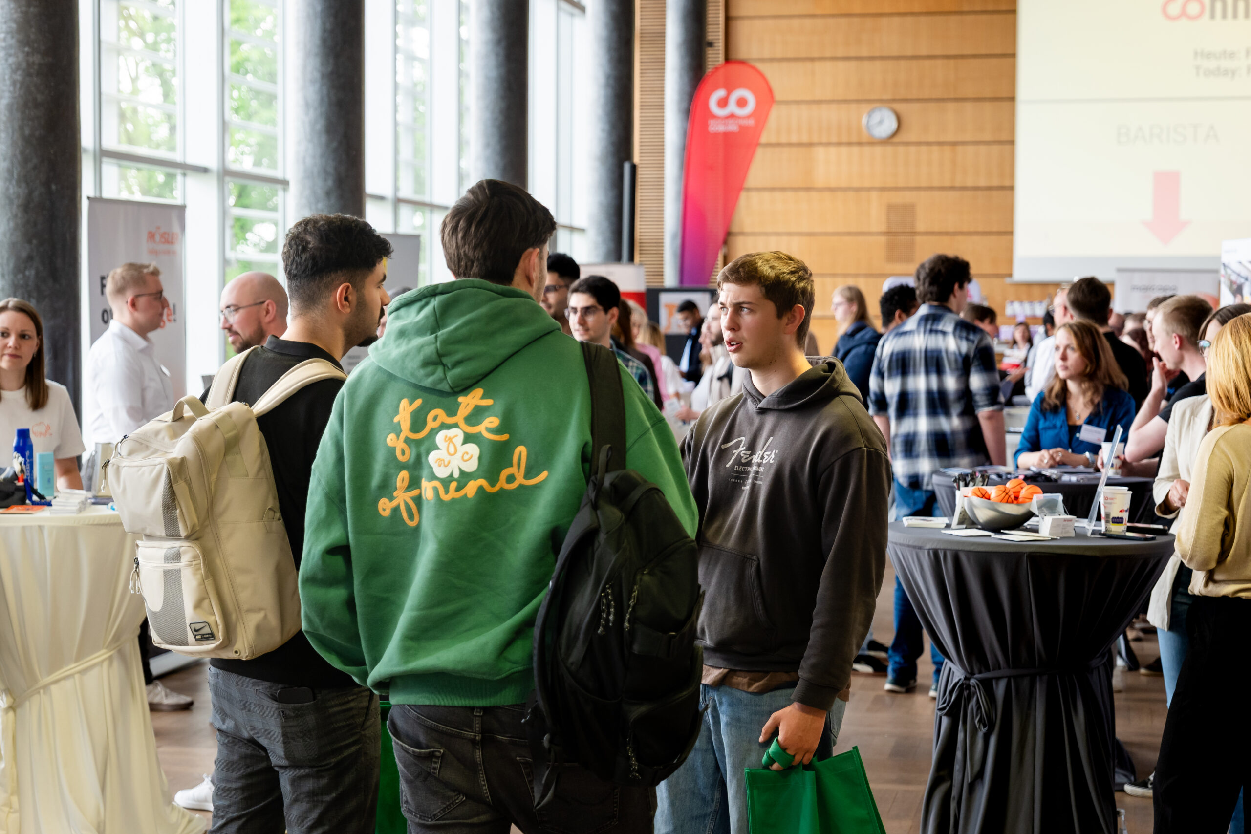 Bei einer Networking-Veranstaltung in einem Innenraum kommen Menschen zusammen und knüpfen Kontakte. Einige stehen an Cocktailtischen, und eine Person trägt einen grünen Kapuzenpulli mit der Aufschrift "state of mind" auf dem Rücken. Die Atmosphäre ist locker und gesellig, ideal für junge Talente, um sich zu treffen und auszutauschen. at Hochschule Coburg