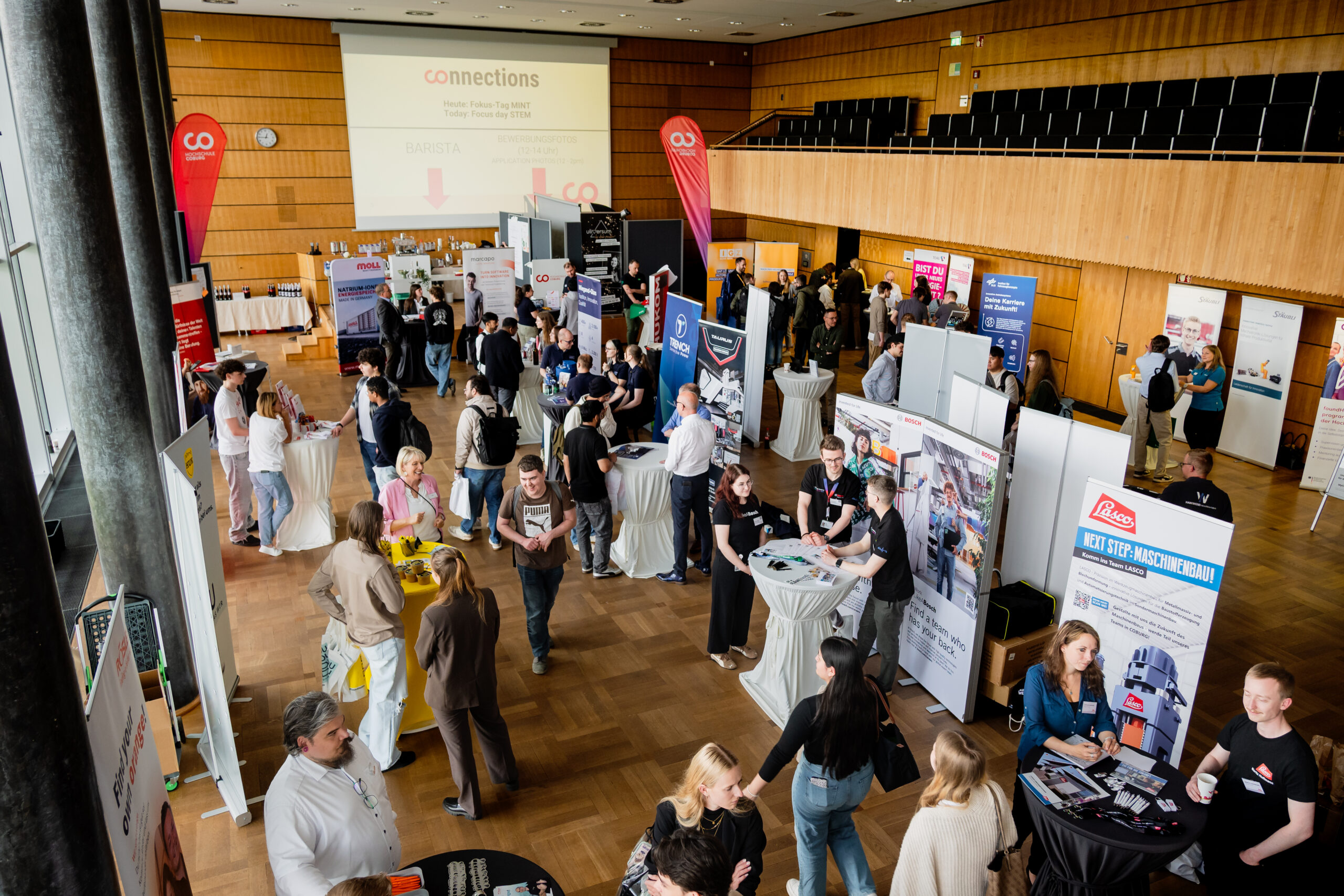Eine belebte Jobmesse oder Networking-Veranstaltung in einer großen Halle, wo Junge Talente an Messeständen mit Fachleuten in Kontakt treten. Auf Bannern und Plakaten sind die Namen der Unternehmen zu lesen, während die Teilnehmer an Stehtischen interagieren und wertvolle Kontakte knüpfen. at Hochschule Coburg