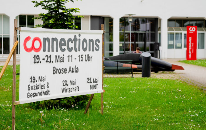 Ein Schild auf einer Wiese wirbt für die Veranstaltung "Connections" vom 19. bis 21. Mai in der Brose Aula, bei der Themen wie soziale Gesundheit, Wirtschaft, MINT und Talente gewinnen im Mittelpunkt stehen. Im Hintergrund sind ein Gebäude und draußen sitzende Menschen zu sehen. at Hochschule Coburg