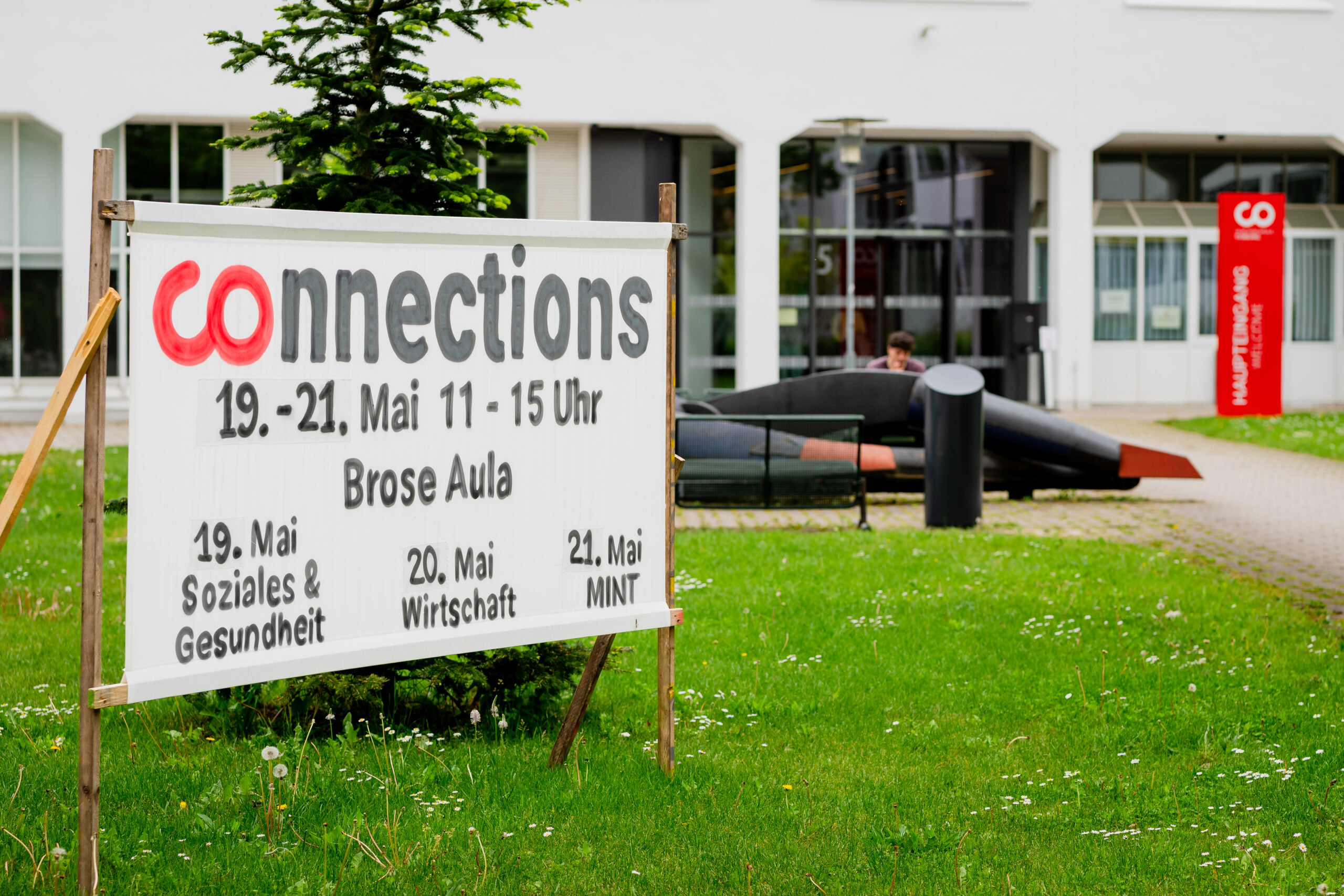 Ein Schild auf einer Wiese wirbt für die Veranstaltung "Connections" vom 19. bis 21. Mai in der Brose Aula, bei der Themen wie soziale Gesundheit, Wirtschaft, MINT und Talente gewinnen im Mittelpunkt stehen. Im Hintergrund sind ein Gebäude und draußen sitzende Menschen zu sehen. at Hochschule Coburg