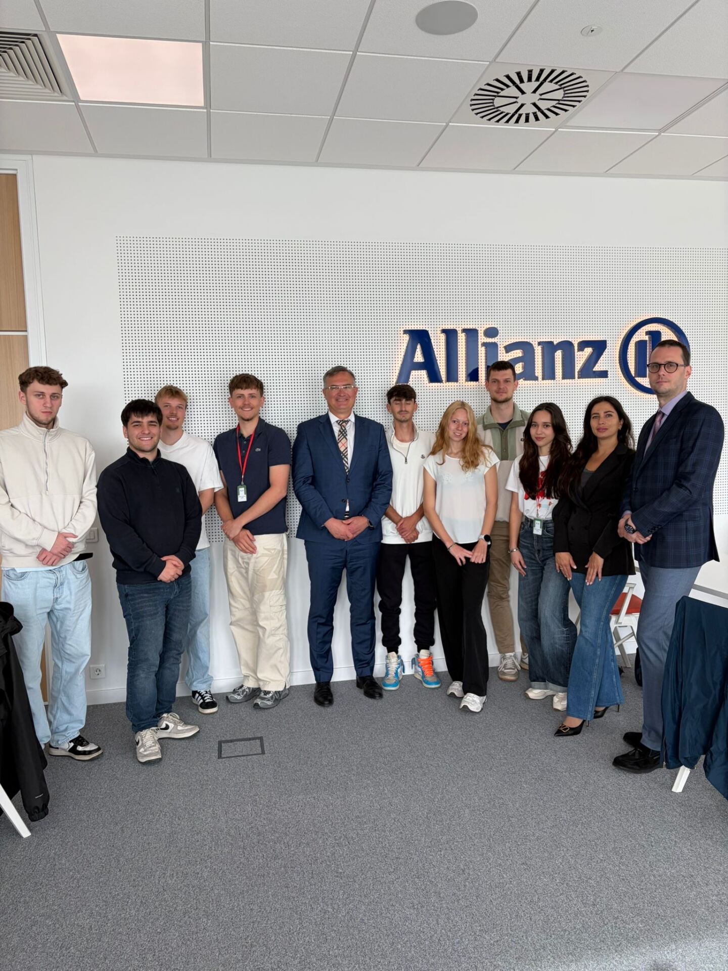 Eine Gruppe von elf Personen, darunter junge Erwachsene und Berufstätige in Geschäftskleidung, steht gemeinsam in einem Büroraum, hinter ihnen ist das Allianz-Logo an der Wand zu sehen, daneben ein „automatisch gespeicherter Entwurf“ auf einem Bildschirm in der Nähe. at Hochschule Coburg