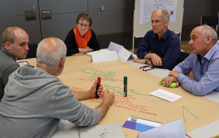 Fünf Erwachsene sitzen um einen Tisch herum, auf dem ein großes Blatt Papier mit handschriftlichen Notizen liegt, und diskutieren und nutzen dabei Filzstifte während einer gemeinsamen Besprechung oder eines Workshops in einer modernen Innenraumumgebung. at Hochschule Coburg
