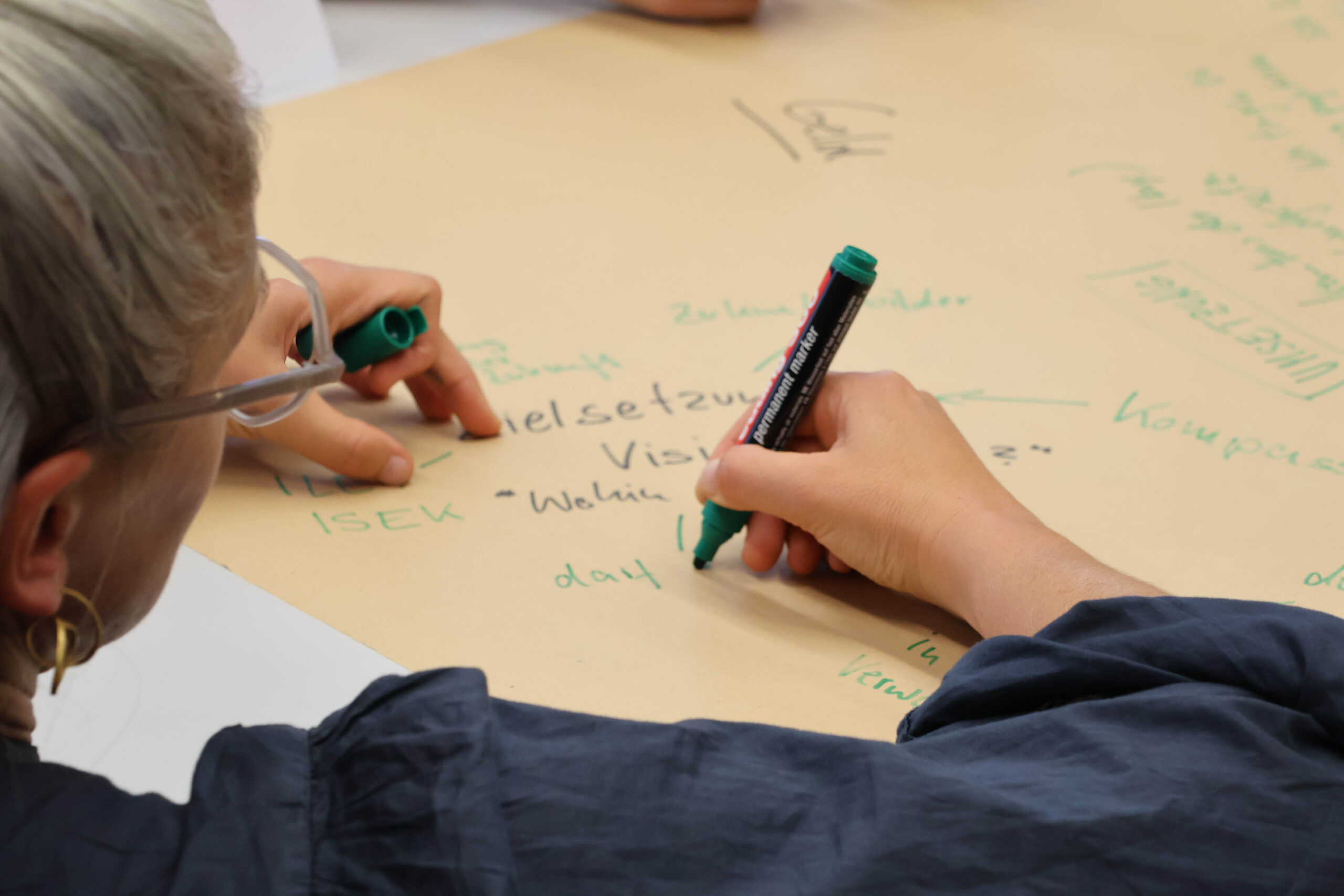 Eine Person mit kurzem grauen Haar und Brille schreibt während einer Gruppen-Brainstorming-Sitzung mit einem grünen Filzstift Notizen auf ein großes Blatt Papier. Daneben ist eine weitere Hand mit einem Filzstift zu sehen. at Hochschule Coburg