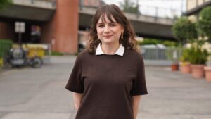 Eine Frau mit welligem braunem Haar, die ein braunes Hemd mit weißem Kragen trägt, steht im Freien und lächelt. Hinter ihr sind Topfpflanzen, ein Fahrrad und eine Überführung zu sehen – eine Szene, die die strahlende Zukunft der Stipendiatinnen widerspiegelt, die sich auf ihre Chancen im Jahr 2026 freuen. at Hochschule Coburg