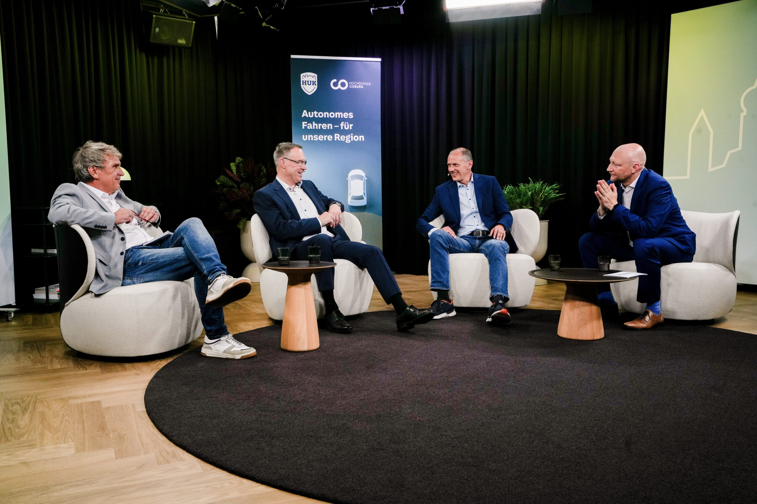 Vier Männer in Geschäftskleidung sitzen in einem Studio auf runden Stühlen und diskutieren. Auf einem Banner hinter ihnen steht „Autonomes Fahren – für unsere Region“. Sie scheinen an einer formellen Podiumsdiskussion oder einem Interview zum Thema autonomes Fahren teilzunehmen, wobei Vertreter der Hochschule Coburg und der HUK-COBURG ihr Fachwissen in die Diskussion einbringen. at Hochschule Coburg