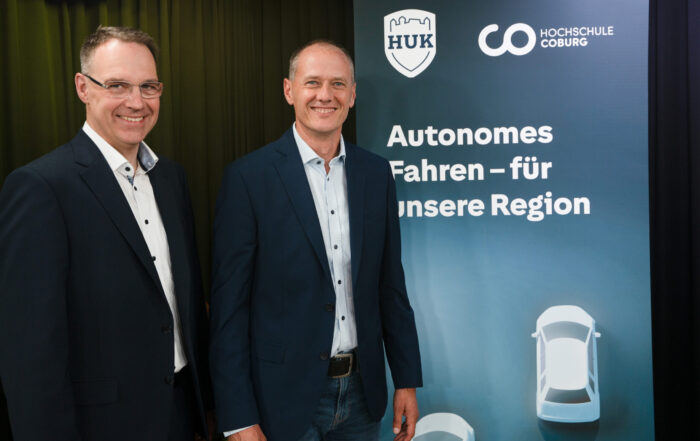 Zwei lächelnde Männer in Anzügen stehen neben einem Schild mit Logos und einem deutschen Text zum Thema „autonomes Fahren“ in der Region. Das Schild, das mit den Logos von HUK-COBURG und der Hochschule Coburg versehen ist, zeigt Autosymbole, die für Fortschritte in der Technologie des autonomen Fahrens stehen. at Hochschule Coburg